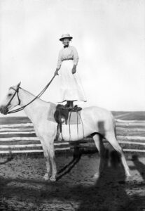 Photographer Evelyn Cameron wearing a hat, smiling, holding reins, and standing on her white horse, Jim. Photo by Evelyn Cameron courtesy of Montana Historical Society Research Center.