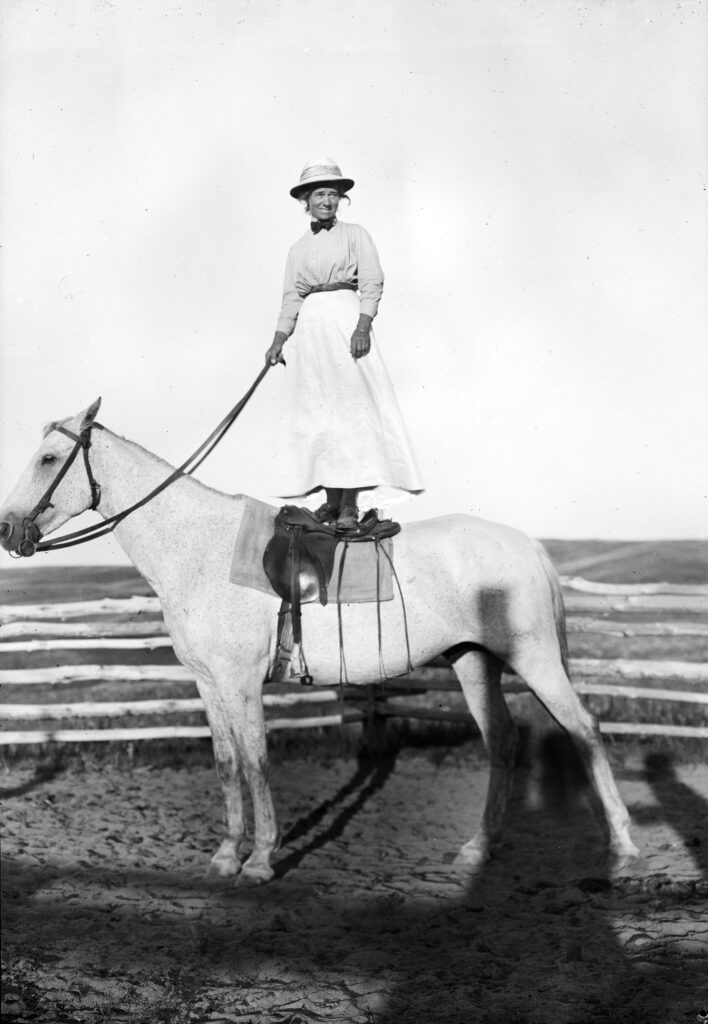 Photographer Evelyn Cameron wearing a hat, smiling, holding reins, and standing on her white horse, Jim. Photo by Evelyn Cameron courtesy of Montana Historical Society Research Center.